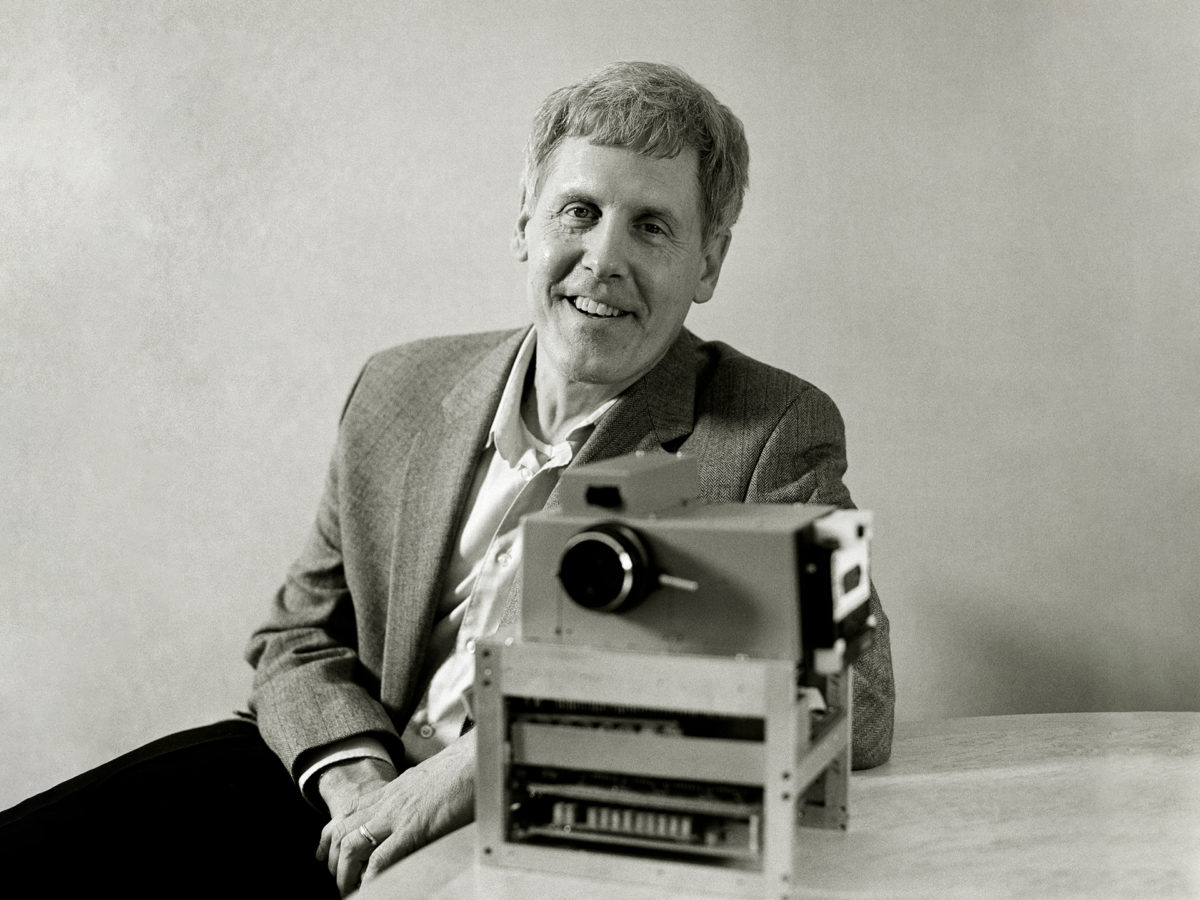 Black-and-white image of a man, Steven Sasson, in black trousers, open-neck business shirt and a light-toned jacket, sitting at desk with his prototype of the world-first digital camera.